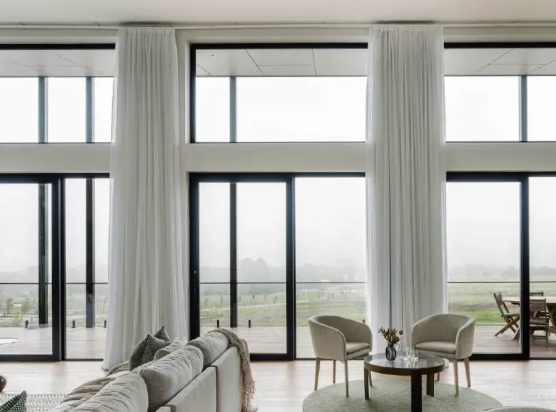 Floor-to-ceiling black-framed windows with sheer curtains overlooking a misty rural view from a living room